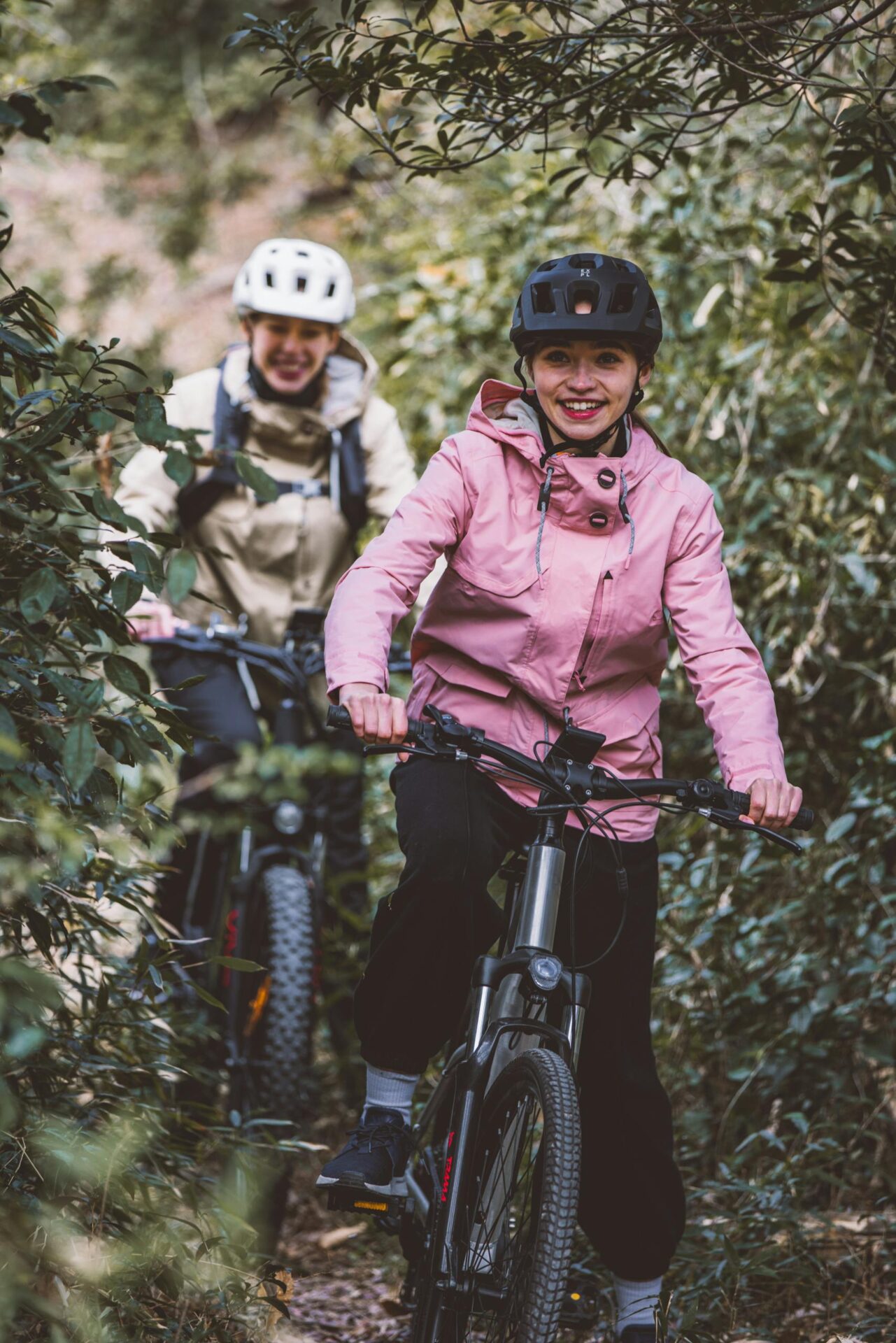 Two women biking on a forest trail, wearing helmets and enjoying the ride outdoors.